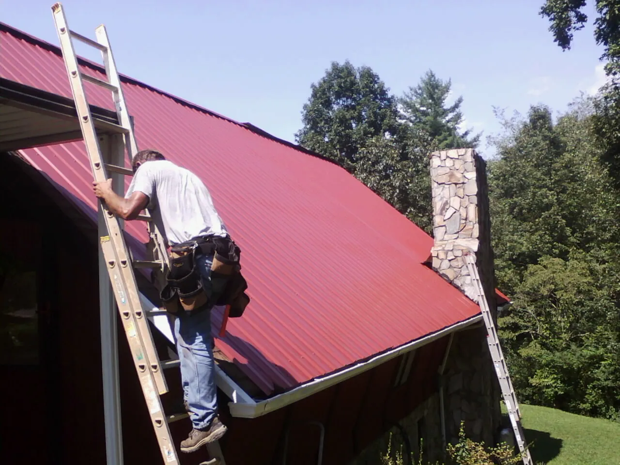 Roofer on a ladder beside a red metal roof and stone chimney — Soffit & Fascia Repair by Roofing Brothers in Duxbury