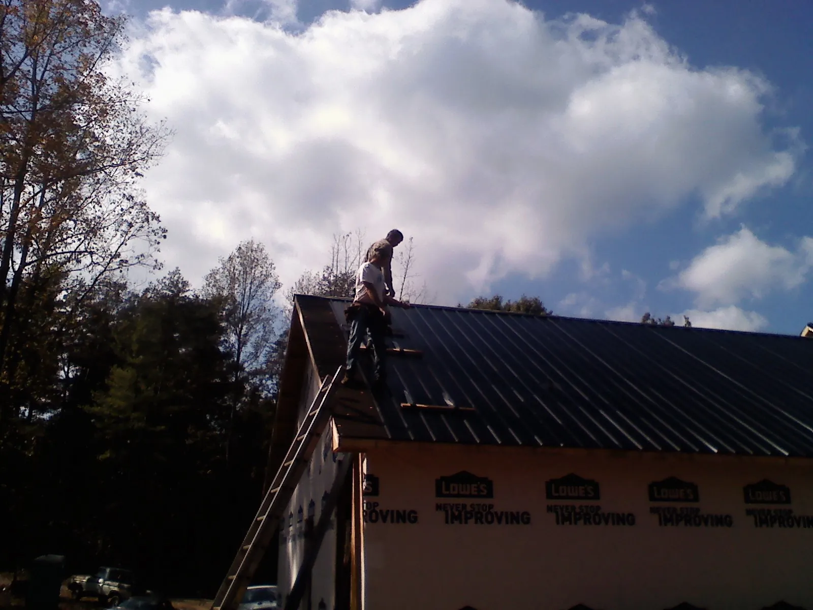 Roofer fastening dark metal panels high against a cloudy sky, from a Roof Inspection job in Duxbury